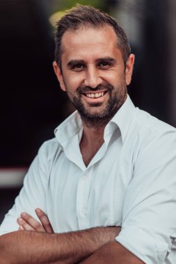 Beyaz gömlekli başarılı bir iş adamı, kollarını kavuşturmuş, açık havada poz veriyor, yüzünde kendinden emin bir ifade var.