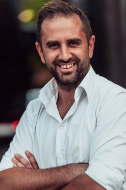 Beyaz gömlekli başarılı bir iş adamı, kollarını kavuşturmuş, açık havada poz veriyor, yüzünde kendinden emin bir ifade var.