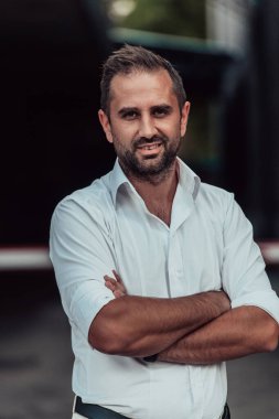 Beyaz gömlekli başarılı bir iş adamı, kollarını kavuşturmuş, açık havada poz veriyor, yüzünde kendinden emin bir ifade var.