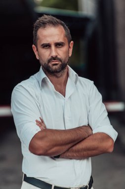 Beyaz gömlekli başarılı bir iş adamı, kollarını kavuşturmuş, açık havada poz veriyor, yüzünde kendinden emin bir ifade var.