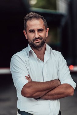 Beyaz gömlekli başarılı bir iş adamı, kollarını kavuşturmuş, açık havada poz veriyor, yüzünde kendinden emin bir ifade var.