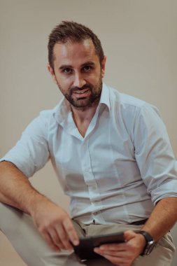 Kendinden emin bir işadamı oturmuş ve kararlı bir ifadeyle tablet kullanırken, bej bir arka plan profesyonel atmosferi yükselterek üretkenliğini ve uzmanlığını gösteriyor.