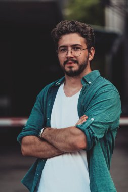 Tişörtlü başarılı genç bir iş adamı, kollarını kavuşturmuş, açık havada poz veriyor, yüzünde kendinden emin bir ifade var.