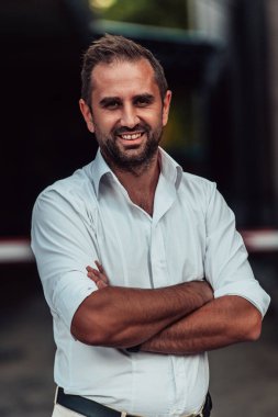 Beyaz gömlekli başarılı bir iş adamı, kollarını kavuşturmuş, açık havada poz veriyor, yüzünde kendinden emin bir ifade var.