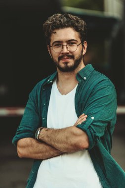 Tişörtlü başarılı genç bir iş adamı, kollarını kavuşturmuş, açık havada poz veriyor, yüzünde kendinden emin bir ifade var.