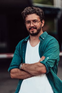 Tişörtlü başarılı genç bir iş adamı, kollarını kavuşturmuş, açık havada poz veriyor, yüzünde kendinden emin bir ifade var.