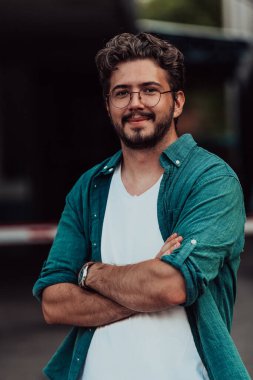 Tişörtlü başarılı genç bir iş adamı, kollarını kavuşturmuş, açık havada poz veriyor, yüzünde kendinden emin bir ifade var.