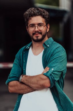Tişörtlü başarılı genç bir iş adamı, kollarını kavuşturmuş, açık havada poz veriyor, yüzünde kendinden emin bir ifade var.