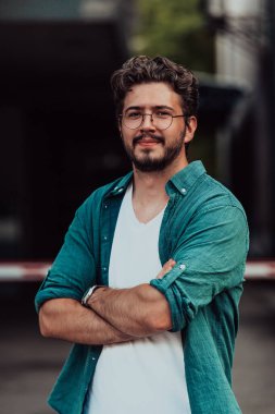 Tişörtlü başarılı genç bir iş adamı, kollarını kavuşturmuş, açık havada poz veriyor, yüzünde kendinden emin bir ifade var.