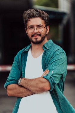 Tişörtlü başarılı genç bir iş adamı, kollarını kavuşturmuş, açık havada poz veriyor, yüzünde kendinden emin bir ifade var.