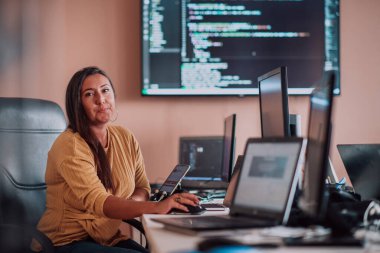 Bilgisayarlarla çevrili bir programcı ofisinde oturan bir iş kadını uzmanlığını ve teknolojiye olan bağlılığını gösteriyor.