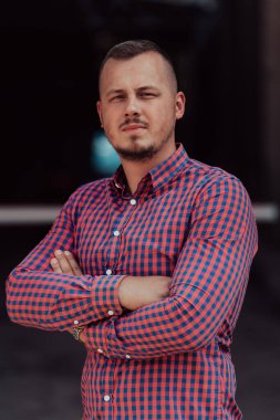 Tişörtlü başarılı genç bir iş adamı, kollarını kavuşturmuş, açık havada poz veriyor, yüzünde kendinden emin bir ifade var.