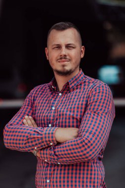 Tişörtlü başarılı genç bir iş adamı, kollarını kavuşturmuş, açık havada poz veriyor, yüzünde kendinden emin bir ifade var.