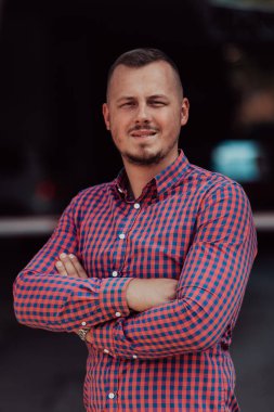 Tişörtlü başarılı genç bir iş adamı, kollarını kavuşturmuş, açık havada poz veriyor, yüzünde kendinden emin bir ifade var.