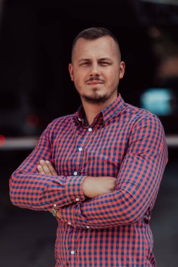 Tişörtlü başarılı genç bir iş adamı, kollarını kavuşturmuş, açık havada poz veriyor, yüzünde kendinden emin bir ifade var.