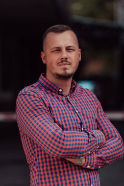 Tişörtlü başarılı genç bir iş adamı, kollarını kavuşturmuş, açık havada poz veriyor, yüzünde kendinden emin bir ifade var.