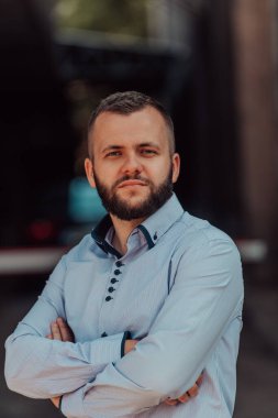 Tişörtlü başarılı genç bir iş adamı, kollarını kavuşturmuş, açık havada poz veriyor, yüzünde kendinden emin bir ifade var.