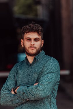 Tişörtlü başarılı genç bir iş adamı, kollarını kavuşturmuş, açık havada poz veriyor, yüzünde kendinden emin bir ifade var.