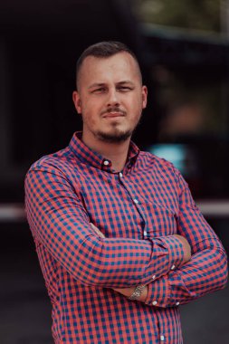 Tişörtlü başarılı genç bir iş adamı, kollarını kavuşturmuş, açık havada poz veriyor, yüzünde kendinden emin bir ifade var.