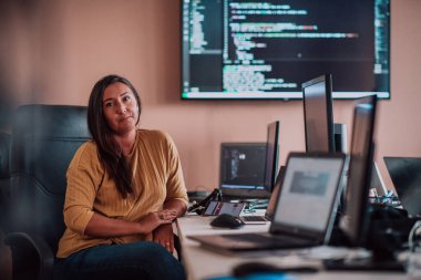 Bilgisayarlarla çevrili bir programcı ofisinde oturan bir iş kadını uzmanlığını ve teknolojiye olan bağlılığını gösteriyor.