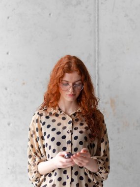 Büyüleyici turuncu saçlı genç bir girişimci şık gri bir duvarın önünde akıllı telefonu tutarken kendine güvenen bir poz veriyor.