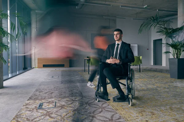 一位坐在现代办公室轮椅上的商人在同事们的簇拥着下 他们的动作模糊不清 象征着他们在工作空间中的支持和团结 — 图库照片
