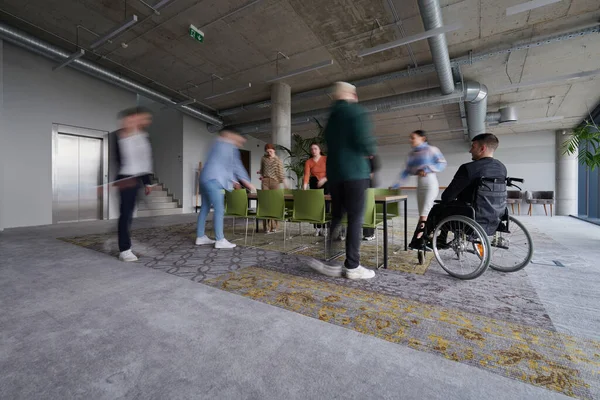 一位坐在轮椅上的商人坐在一个现代化的大办公室里的桌子旁 而他的同事们则聚首一堂 步履维艰 象征着面对挑战时的包容性 支持和团结 — 图库照片