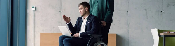 在一个时髦的办公室里 坐在轮椅上的商人身后使用笔记本电脑的全景照片是他的同事给他的支持 — 图库照片