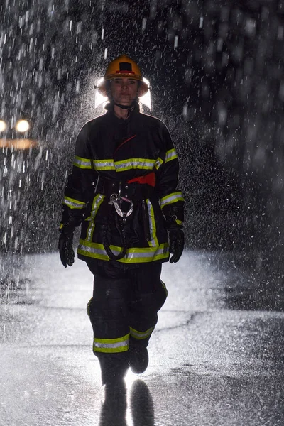 一名身穿职业制服的坚定的女消防员在险象环生的雨夜中大步走过 勇敢地执行营救任务 表现出她坚定不移的勇气和拯救生命的决心 — 图库照片