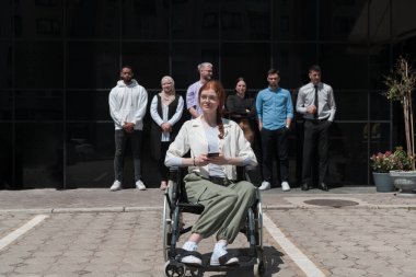 Çeşitli ve kendine güvenen bir grup genç iş adamı birlikte poz veriyor. Tekerlekli sandalyedeki iş kadını başarı, hırs ve birlik saçarak dinamik ve ilham verici bir işin özünü yakalıyor.