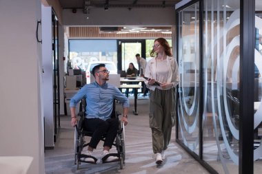 Genç iş arkadaşları, işbirlikçi iş arkadaşları, aralarında tekerlekli sandalyedeki bir kişinin de bulunduğu, modern cam ofis koridorunun yanından geçerek, çeşitliliği, takım çalışmasını ve güçlendirmeyi resmediyorlar.