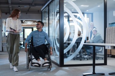 Genç iş arkadaşları, işbirlikçi iş arkadaşları, aralarında tekerlekli sandalyedeki bir kişinin de bulunduğu, modern cam ofis koridorunun yanından geçerek, çeşitliliği, takım çalışmasını ve güçlendirmeyi resmediyorlar.