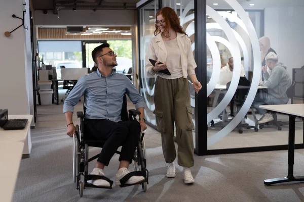 stock image Young business colleagues, collaborative business colleagues, including a person in a wheelchair, walk past a modern glass office corridor, illustrating diversity, teamwork and empowerment in the