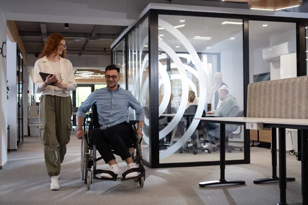 stock image Young business colleagues, collaborative business colleagues, including a person in a wheelchair, walk past a modern glass office corridor, illustrating diversity, teamwork and empowerment in the