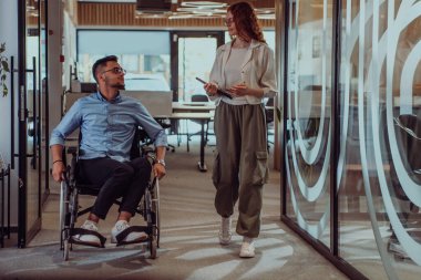 Genç iş arkadaşları, işbirlikçi iş arkadaşları, aralarında tekerlekli sandalyedeki bir kişinin de bulunduğu, modern cam ofis koridorunun yanından geçerek, çeşitliliği, takım çalışmasını ve güçlendirmeyi resmediyorlar.