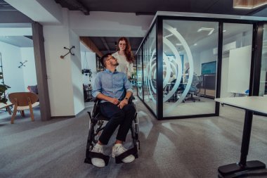 Genç iş arkadaşları, işbirlikçi iş arkadaşları, aralarında tekerlekli sandalyedeki bir kişinin de bulunduğu, modern cam ofis koridorunun yanından geçerek, çeşitliliği, takım çalışmasını ve güçlendirmeyi resmediyorlar.