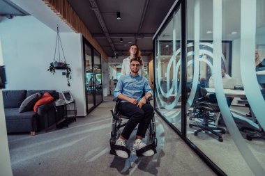 Genç iş arkadaşları, işbirlikçi iş arkadaşları, aralarında tekerlekli sandalyedeki bir kişinin de bulunduğu, modern cam ofis koridorunun yanından geçerek, çeşitliliği, takım çalışmasını ve güçlendirmeyi resmediyorlar.