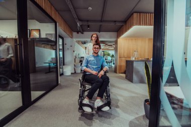 Genç iş arkadaşları, işbirlikçi iş arkadaşları, aralarında tekerlekli sandalyedeki bir kişinin de bulunduğu, modern cam ofis koridorunun yanından geçerek, çeşitliliği, takım çalışmasını ve güçlendirmeyi resmediyorlar.