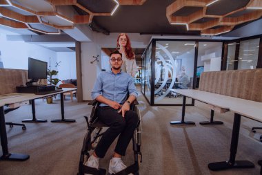 Genç iş arkadaşları, işbirlikçi iş arkadaşları, aralarında tekerlekli sandalyedeki bir kişinin de bulunduğu, modern cam ofis koridorunun yanından geçerek, çeşitliliği, takım çalışmasını ve güçlendirmeyi resmediyorlar.