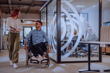 Genç iş arkadaşları, işbirlikçi iş arkadaşları, aralarında tekerlekli sandalyedeki bir kişinin de bulunduğu, modern cam ofis koridorunun yanından geçerek, çeşitliliği, takım çalışmasını ve güçlendirmeyi resmediyorlar.