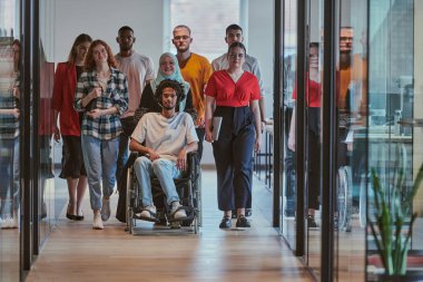 Modern bir girişimin cam kaplı ofisinde koridorda yürüyen bir grup genç iş adamı arasında tekerlekli sandalyedeki bir kişi ve tesettürlü bir kadın da var.