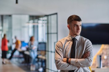 Genç bir iş adamı modern bir ofis koridorunda kollarını kavuşturarak duruyor. Kendine güven ve bir amaç duygusu yayıyor. Dinamik ve ilham verici bir varlığı somutlaştırıyor.