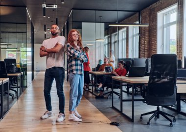 Genç bir Afrikalı Amerikalı işadamı ve turuncu saçlı modern bir iş kadını kollarını kavuşturmuş, çağdaş ofis ortamında güven ve birlik saçan, dinamiği simgeleyen