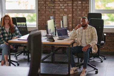 Bir grup iş arkadaşı, canlı turuncu saçlı bir kadın ve genç bir Afro-Amerikan iş adamı, modern bir ofis yerinde oturuyor, çeşitli iş birliğini ve dinamik çalışma ortamını simgeliyor.
