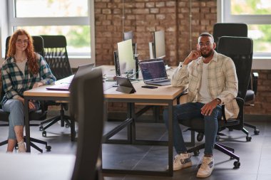 Bir grup iş arkadaşı, canlı turuncu saçlı bir kadın ve genç bir Afro-Amerikan iş adamı, modern bir ofis yerinde oturuyor, çeşitli iş birliğini ve dinamik çalışma ortamını simgeliyor.