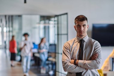 Genç bir iş adamı modern bir ofis koridorunda kollarını kavuşturarak duruyor. Kendine güven ve bir amaç duygusu yayıyor. Dinamik ve ilham verici bir varlığı somutlaştırıyor.