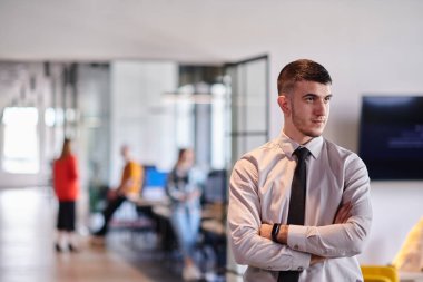 Genç bir iş adamı modern bir ofis koridorunda kollarını kavuşturarak duruyor. Kendine güven ve bir amaç duygusu yayıyor. Dinamik ve ilham verici bir varlığı somutlaştırıyor.