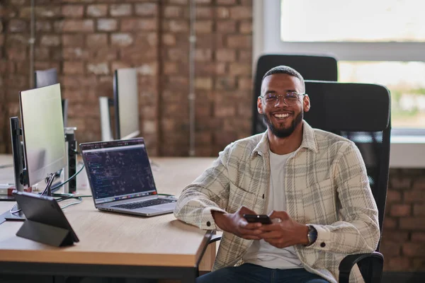 stock image A young African American entrepreneur takes a break in a modern office, using a smartphone to browse social media, capturing a moment of digital connectivity and relaxation amidst his business