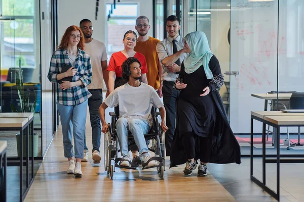 現代のスタートアップのガラス張りのオフィスで廊下を歩く若いビジネスの人々の多様なグループは 車椅子やヒジャブを着用している女性を含み ダイナミックなミックスを示しています — ストック写真
