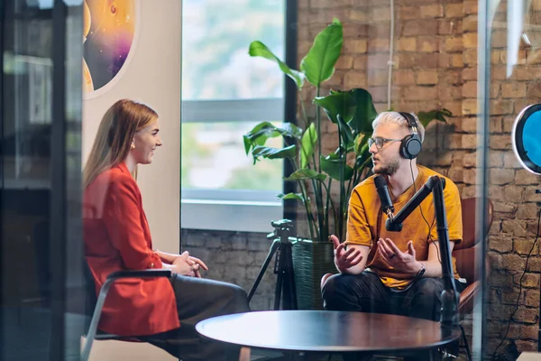 A gathering of young business professionals, some seated in a glass-walled office, engage in a lively conversation and record an online podcast, embodying modern collaboration and dynamic interaction